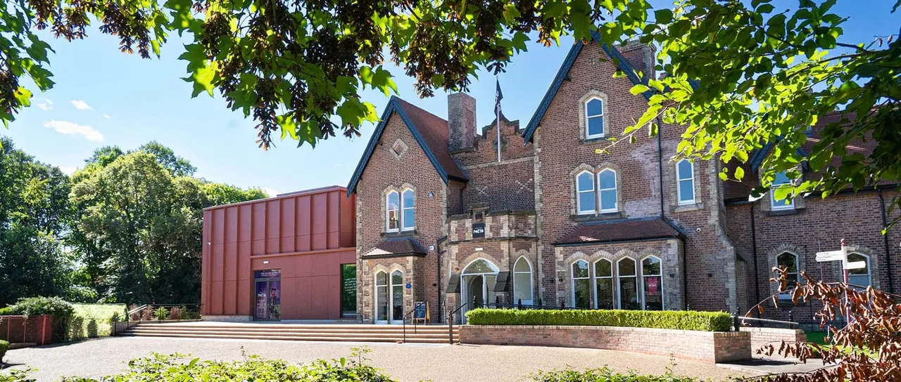 Brick Victorian building with modern red extension, framed by green foliage, on a sunny day.