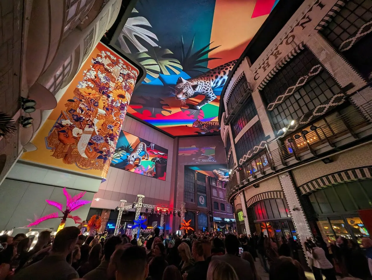 Crowd gathered in an urban venue at night beneath colourful illuminated ceiling panels featuring wildlife imagery and tropical plants.