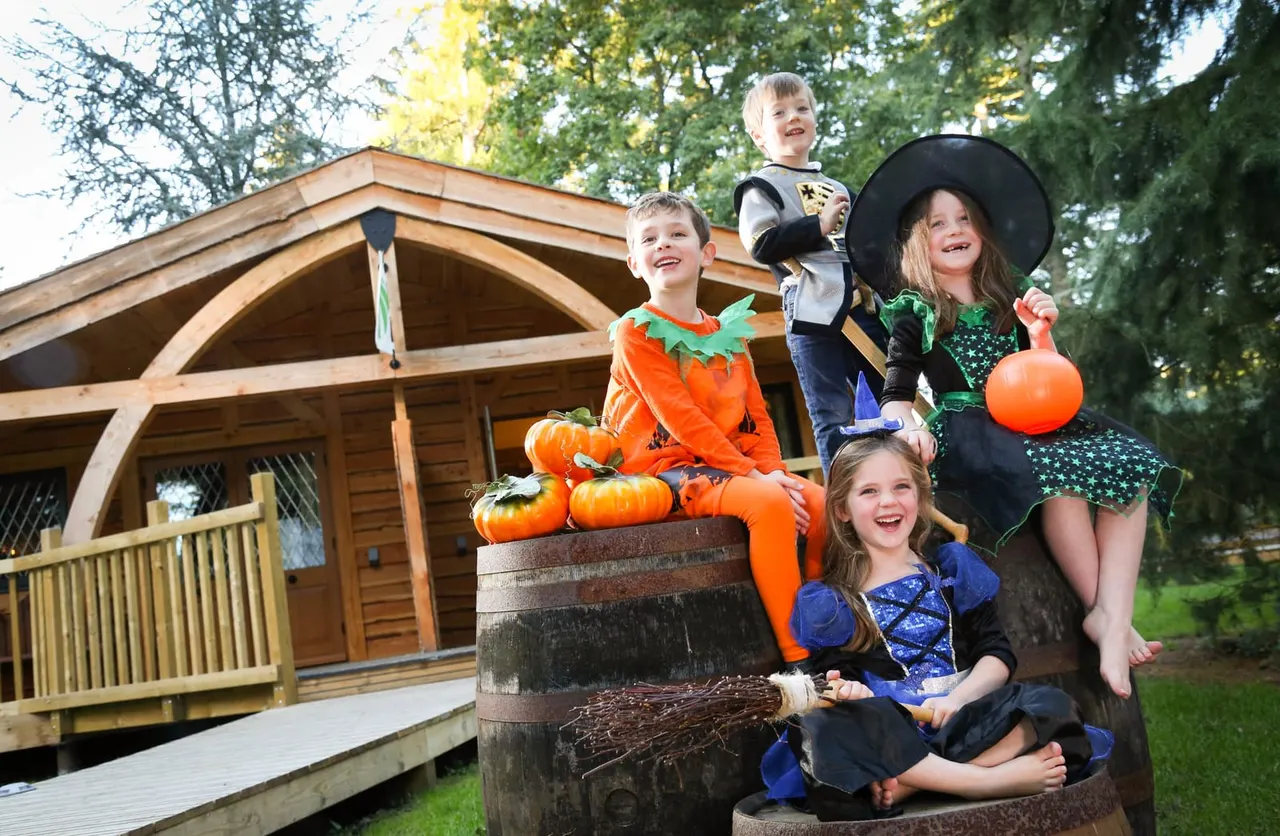 Children in Halloween costumes posing with pumpkins and props outside a wooden lodge building.