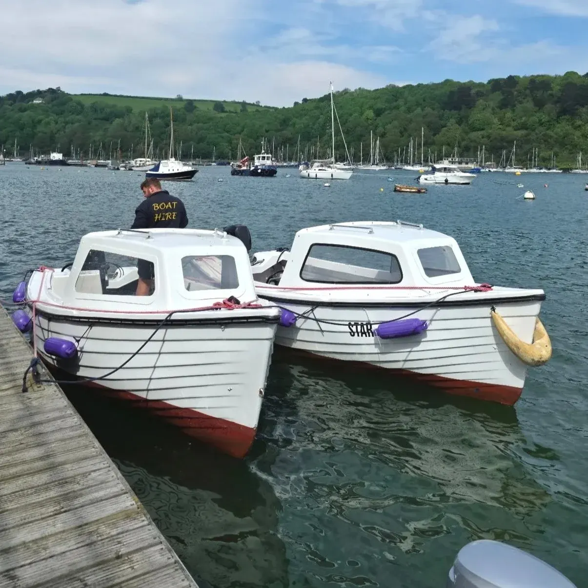 Man in "Boat Hire" jacket stands on two white and red hire boats moored in a river estuary with sailing boats and forested hills beyond.