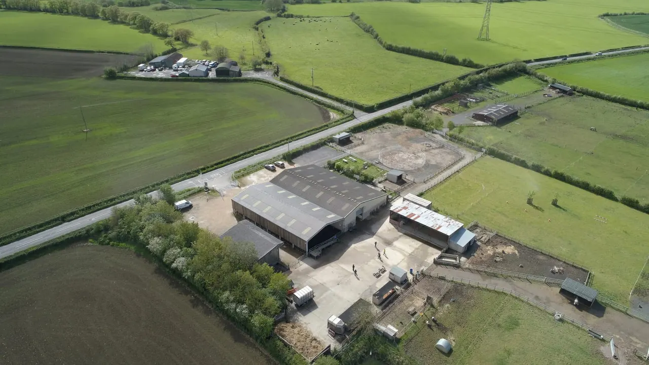 Aerial view of a rural farm complex with large barn buildings, storage structures, and paved courtyard surrounded by green fields and hedgerows.