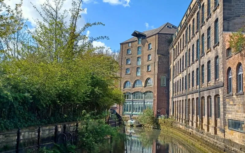 Canal-side view of brick mill buildings reflected in still water, with green foliage and trees flanking the waterway.