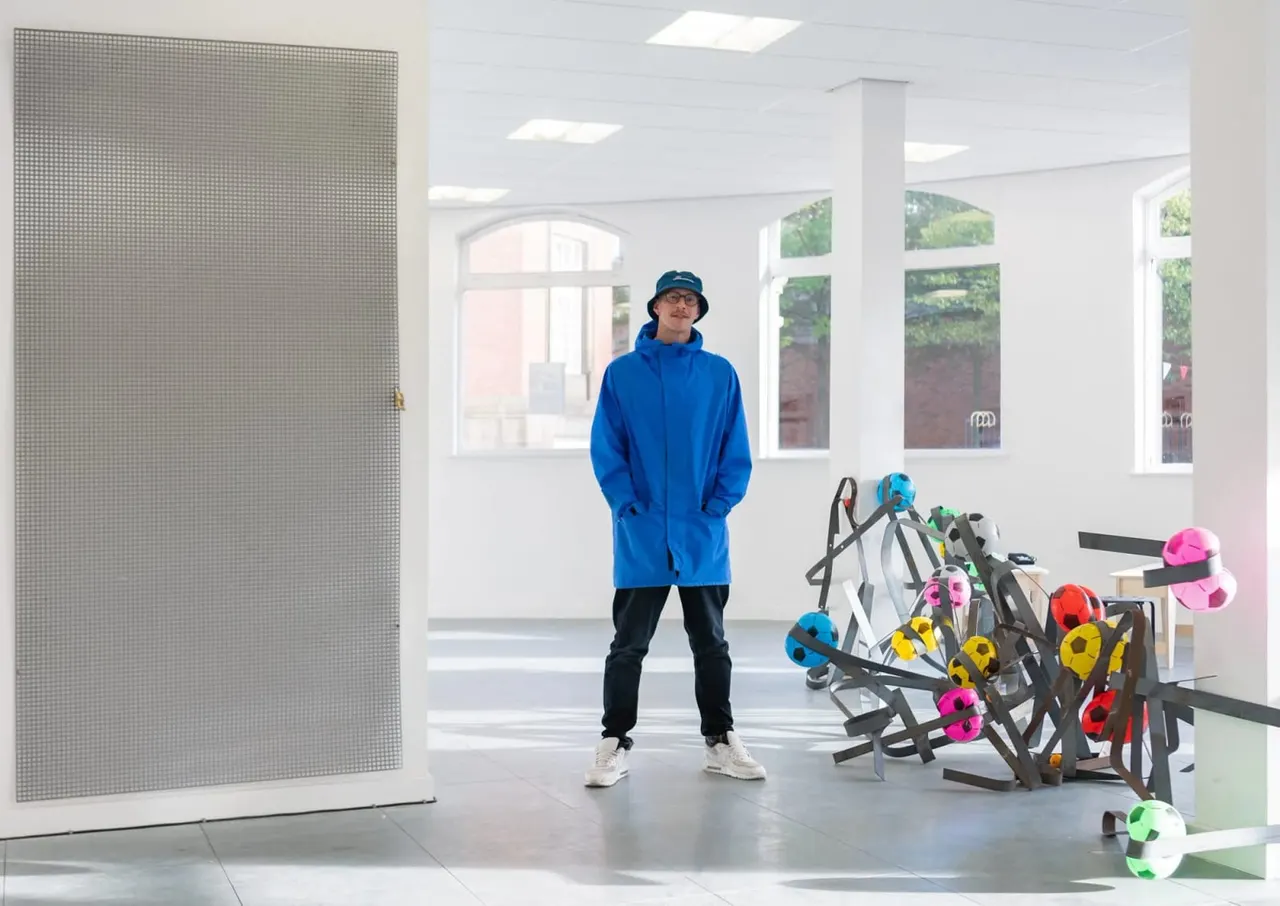 A person in a blue jacket and hat stands in a bright indoor space next to a collection of colourful helmets and sports equipment.