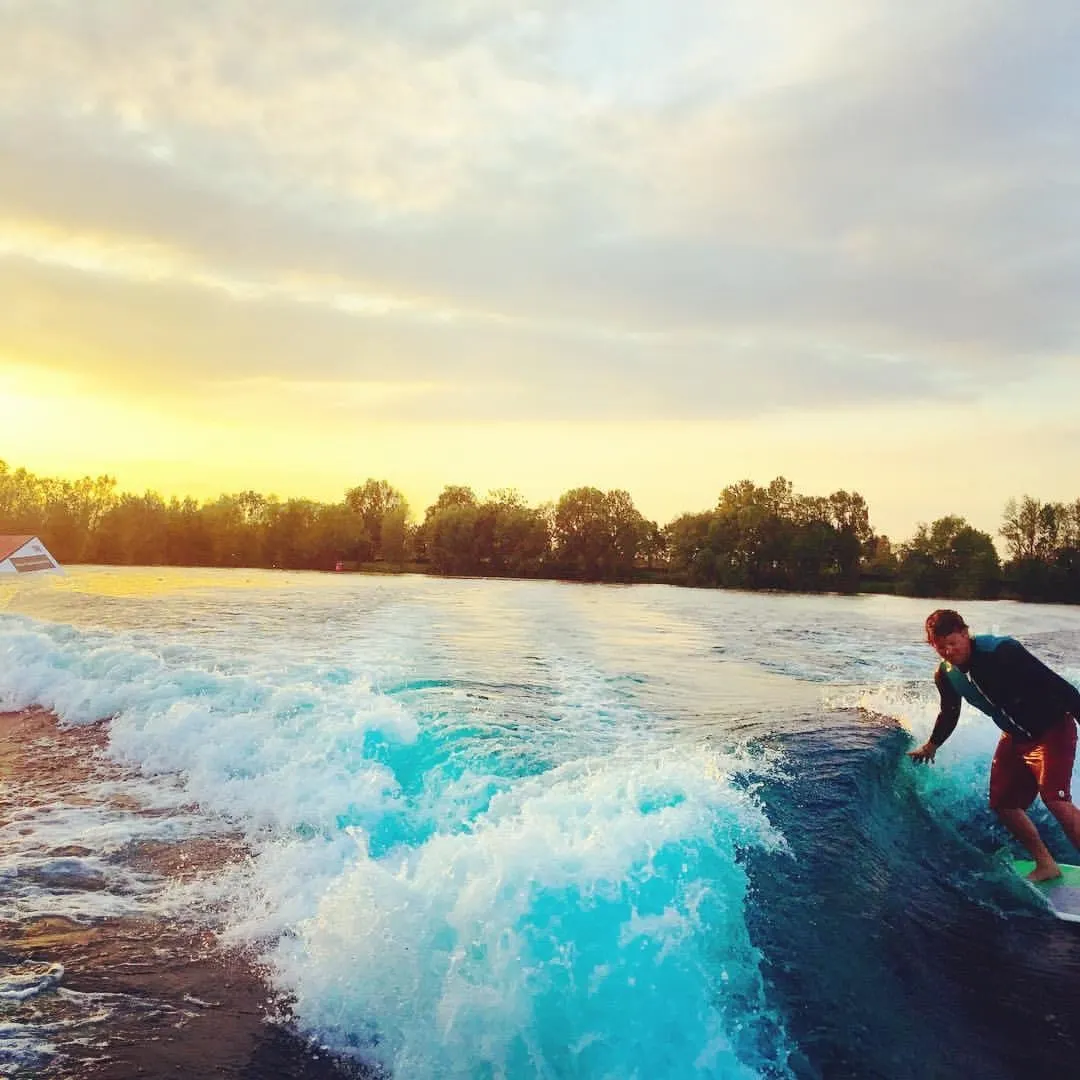 Person surfing on an artificial wave at sunset with trees and lake in the background.
