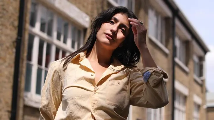 Woman in beige shirt standing outdoors against a brick building, gazing upward.
