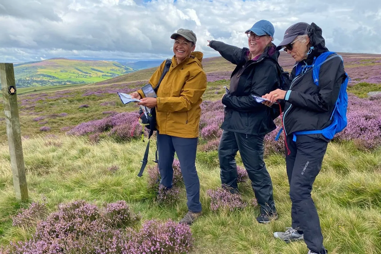 Three hikers consult a map on a moorland hillside covered in purple heather and flowering vegetation.