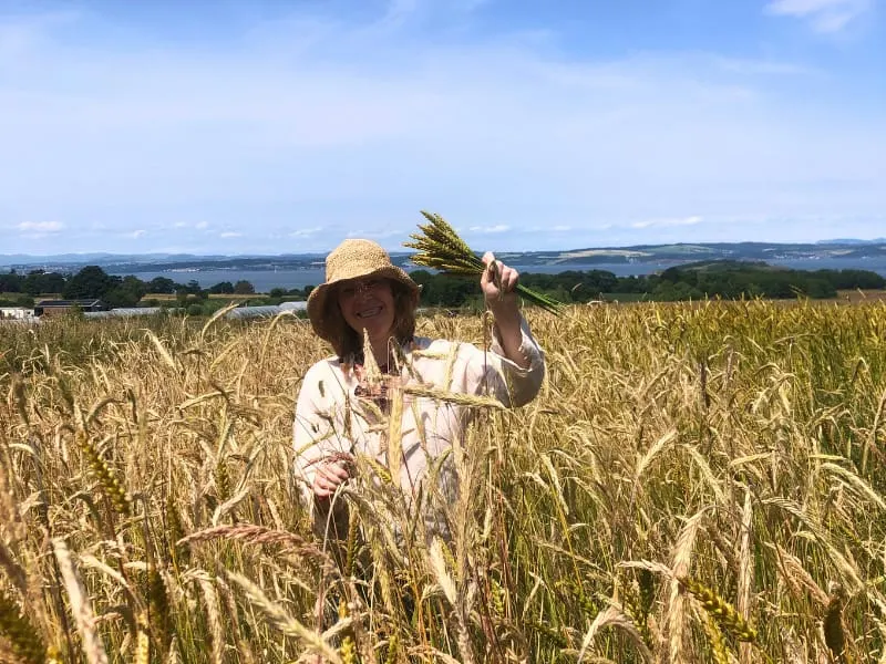 Woman in straw hat holding grain in a golden cereal field with countryside views beyond.