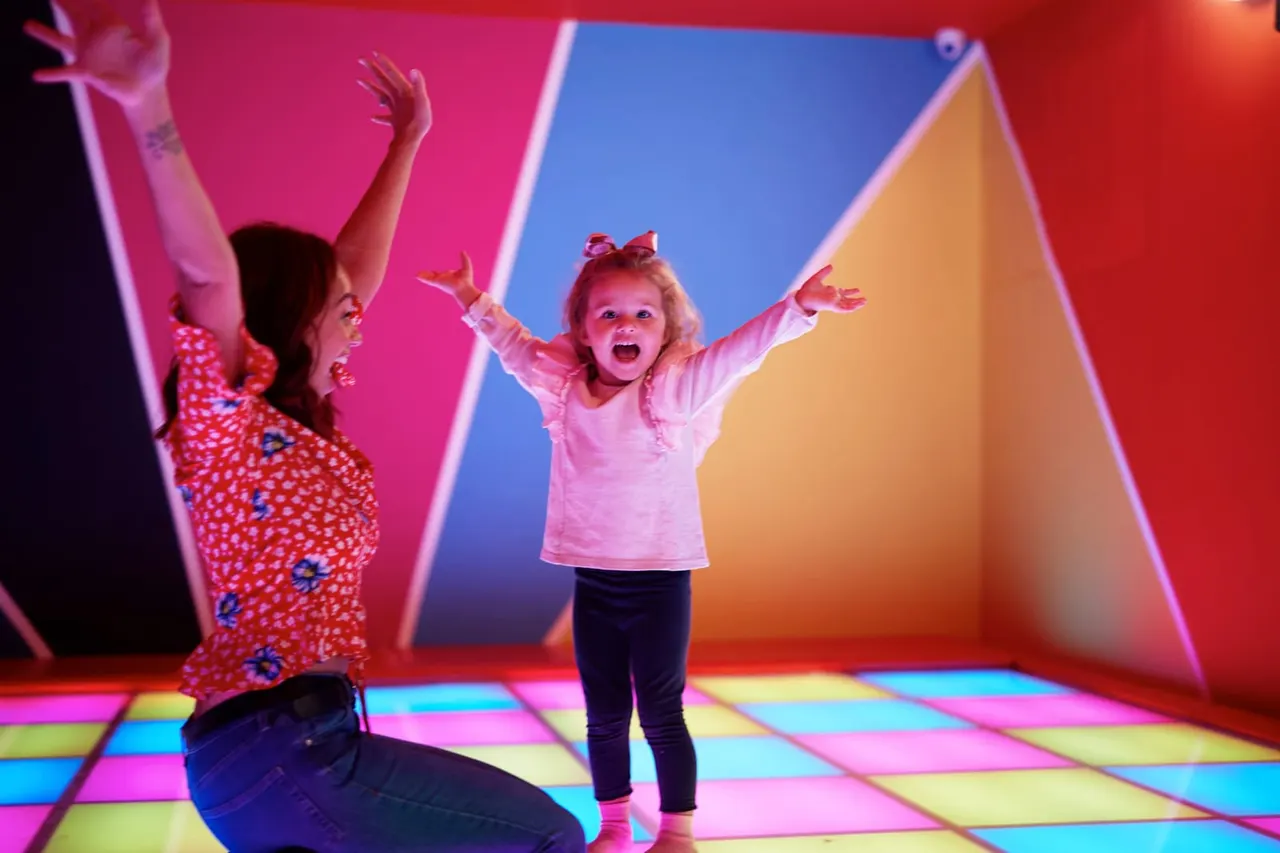 Two young children playing in a brightly lit indoor space with colourful neon walls and a rainbow tile floor.