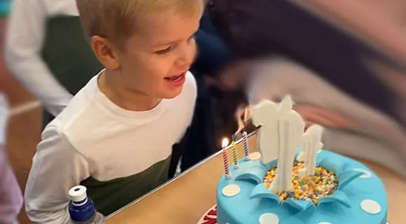 Young child in white and green top blowing out candles on a blue birthday cake.