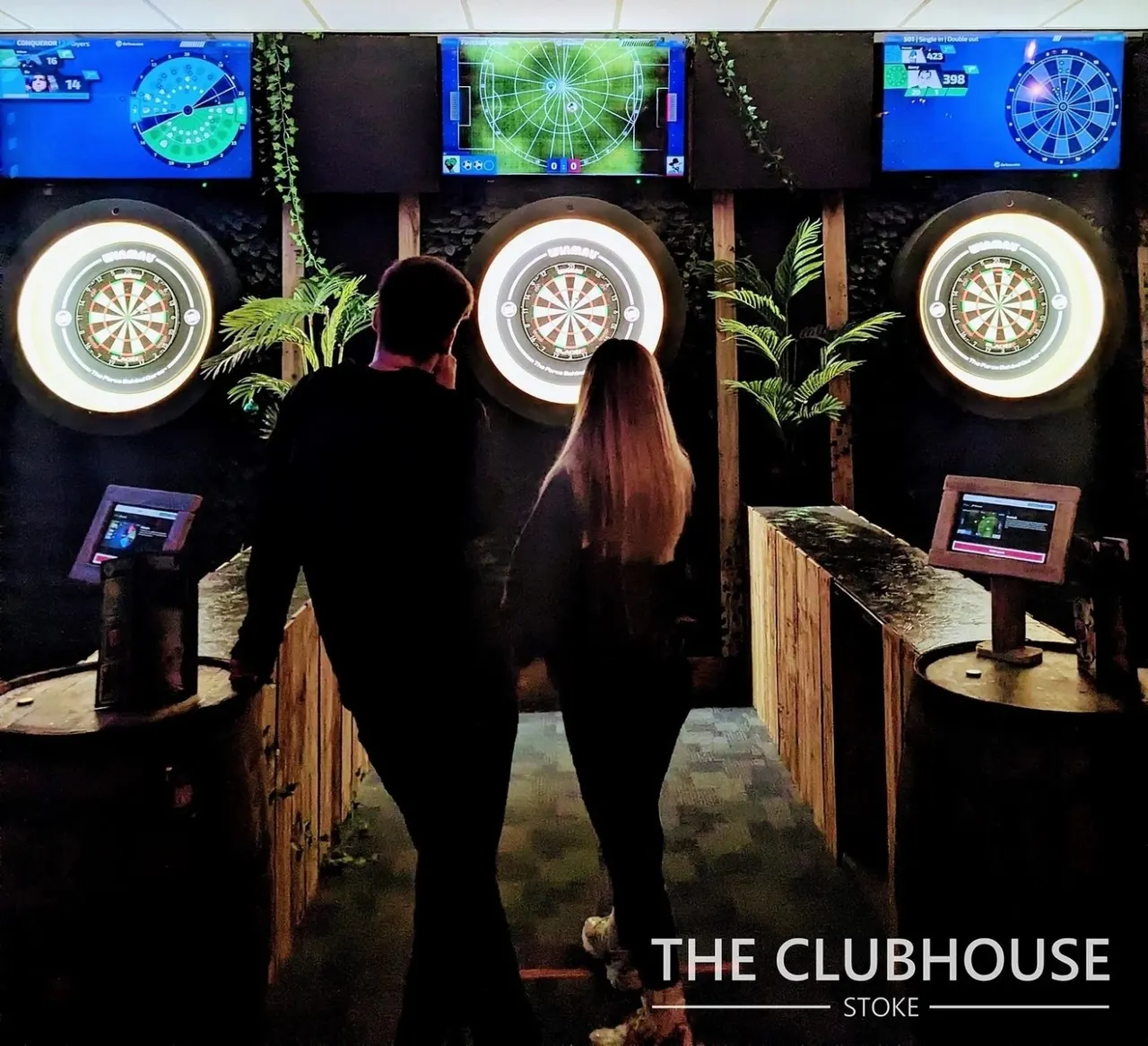 Two people play darts at an illuminated electronic dartboard in a modern leisure venue with screens above.