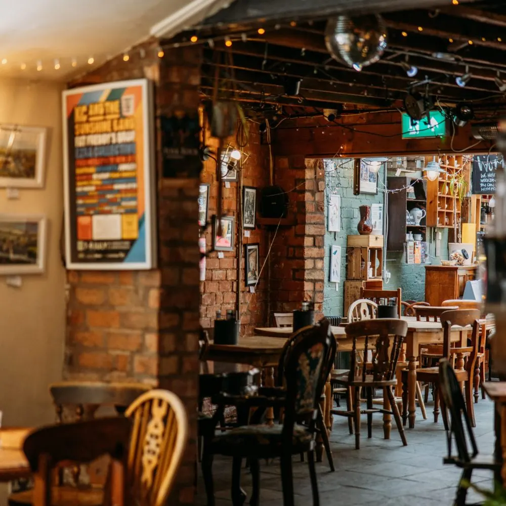 A rustic cafe interior with wooden tables and chairs, brick walls decorated with posters and artwork, warm lighting, and shelving displaying vintage items.