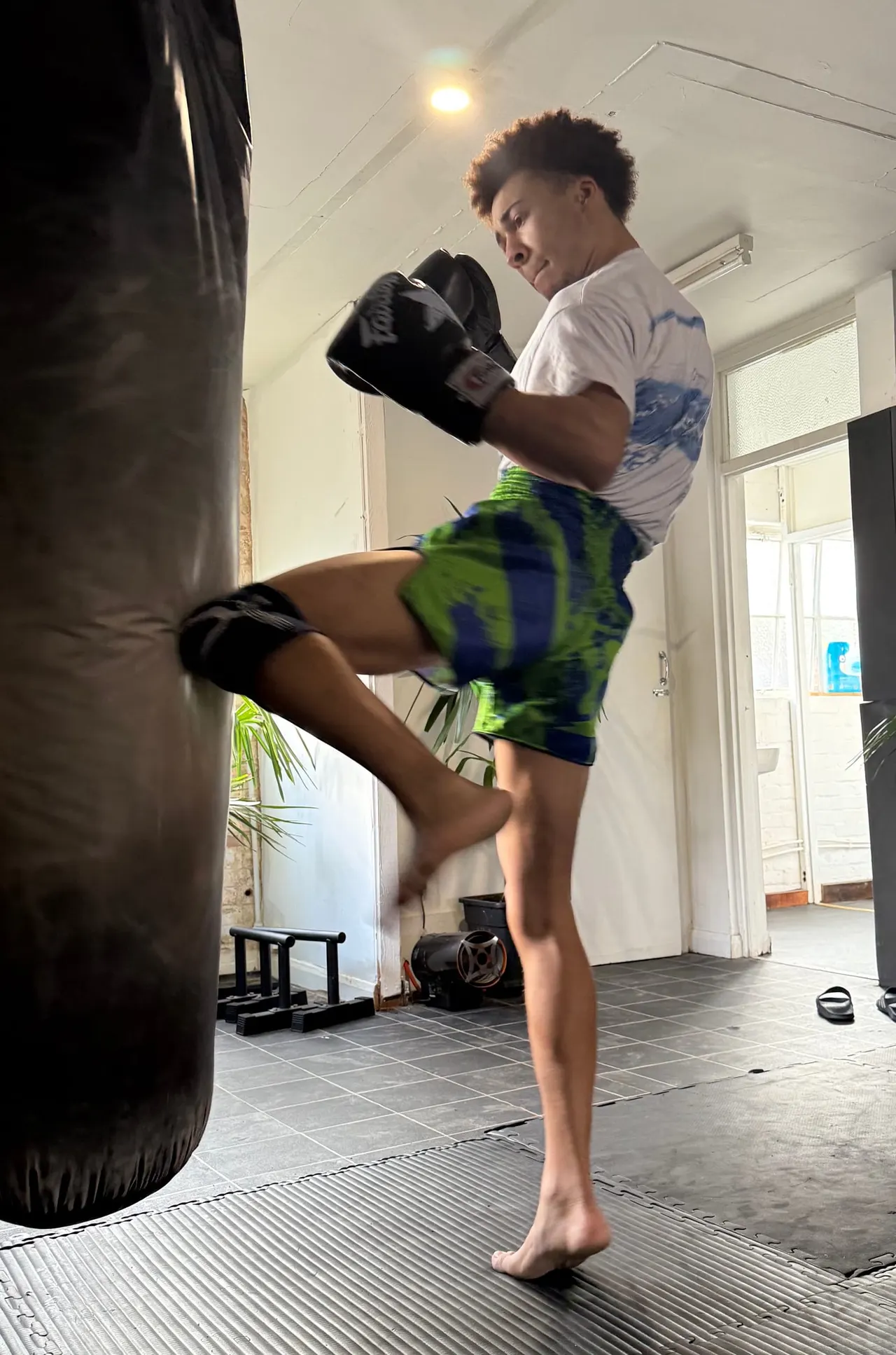 Young person in boxing gloves performing a high kick at a heavy bag in an indoor gym studio.