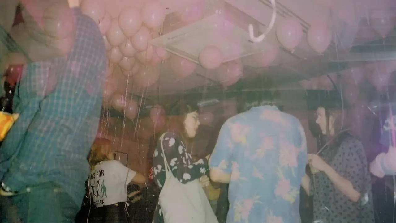 A group of people dancing indoors under pink balloons in a hazy, atmospheric setting.