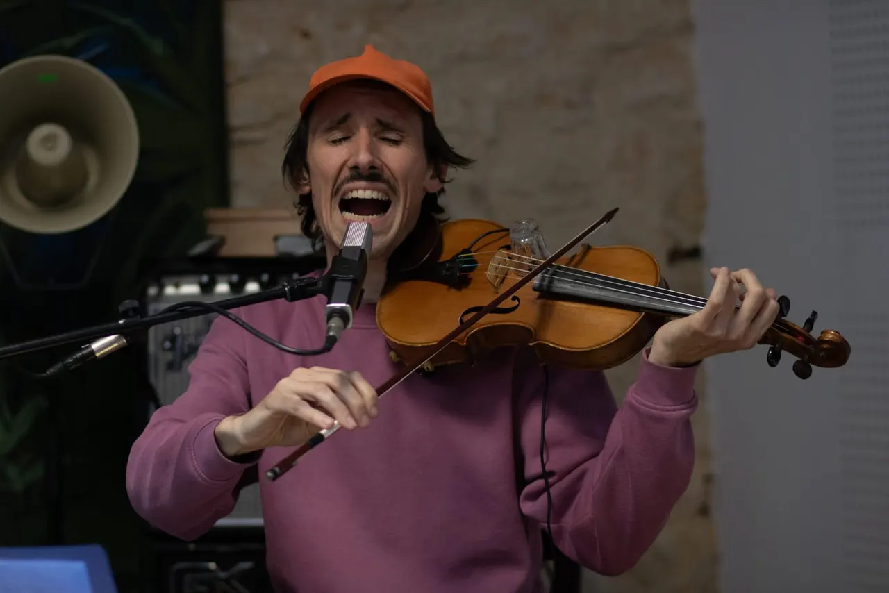 A man in an orange cap and purple sweatshirt sings into a microphone whilst playing a violin during an indoor performance.