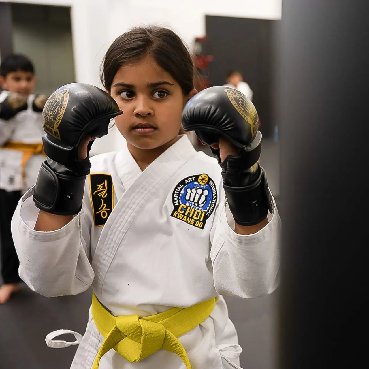 Young girl in white martial arts uniform with yellow belt and black boxing gloves in fighting stance.