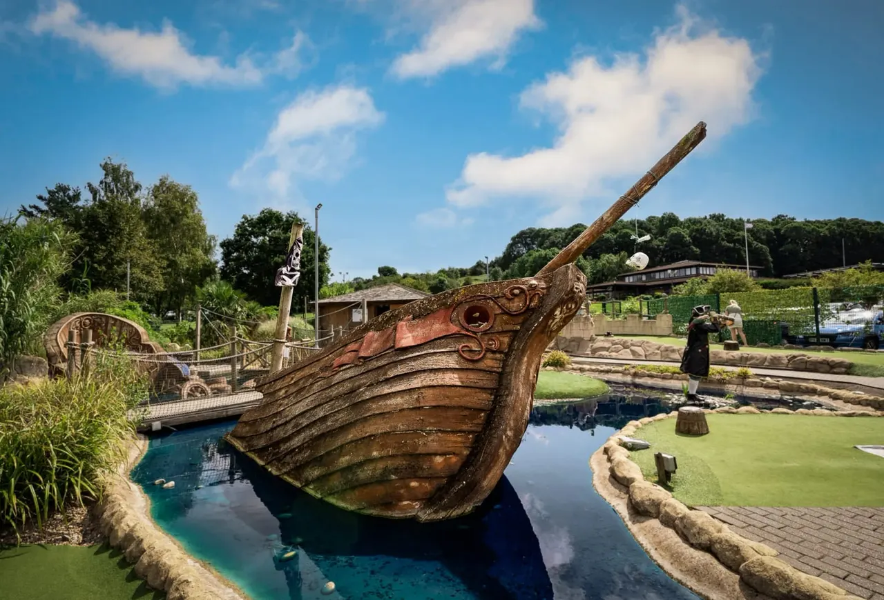 A wooden pirate ship figurehead on a mini golf course with a water channel, visitors, and trees visible in the background.