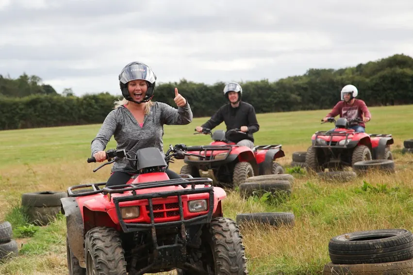 Three people riding red quad bikes on a grass field with tyres as obstacles, wearing helmets.