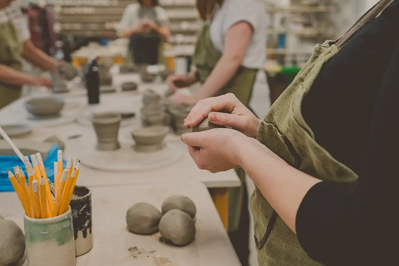 Child's hands shaping clay at a pottery wheel during a ceramics workshop with other participants and materials visible.