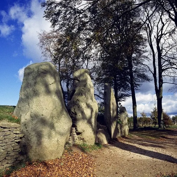 Ancient stone megalith monument with tall standing stones and trees under blue sky.