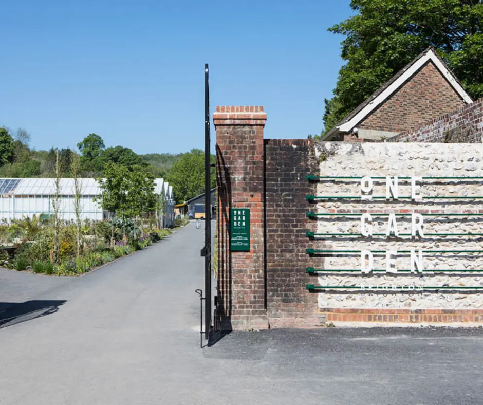 Entrance gate to a country estate with brick pillars, white and green railings, and tree-lined grounds on a sunny day.