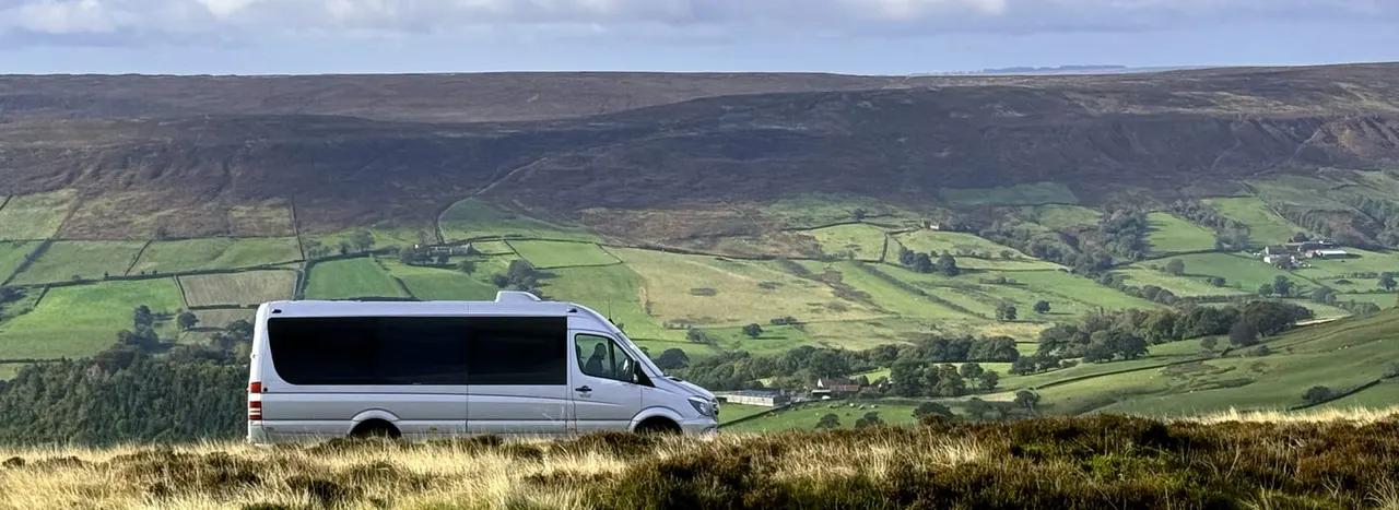 Silver minibus parked on moorland overlooking a patchwork valley of green fields and scattered farmhouses.