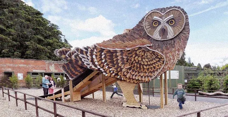 Giant wooden owl sculpture with visitors exploring around it in an outdoor play area.