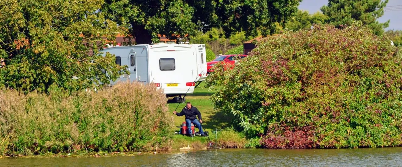 Person on red bike parked by water's edge beside caravan and cars at lakeside campsite.