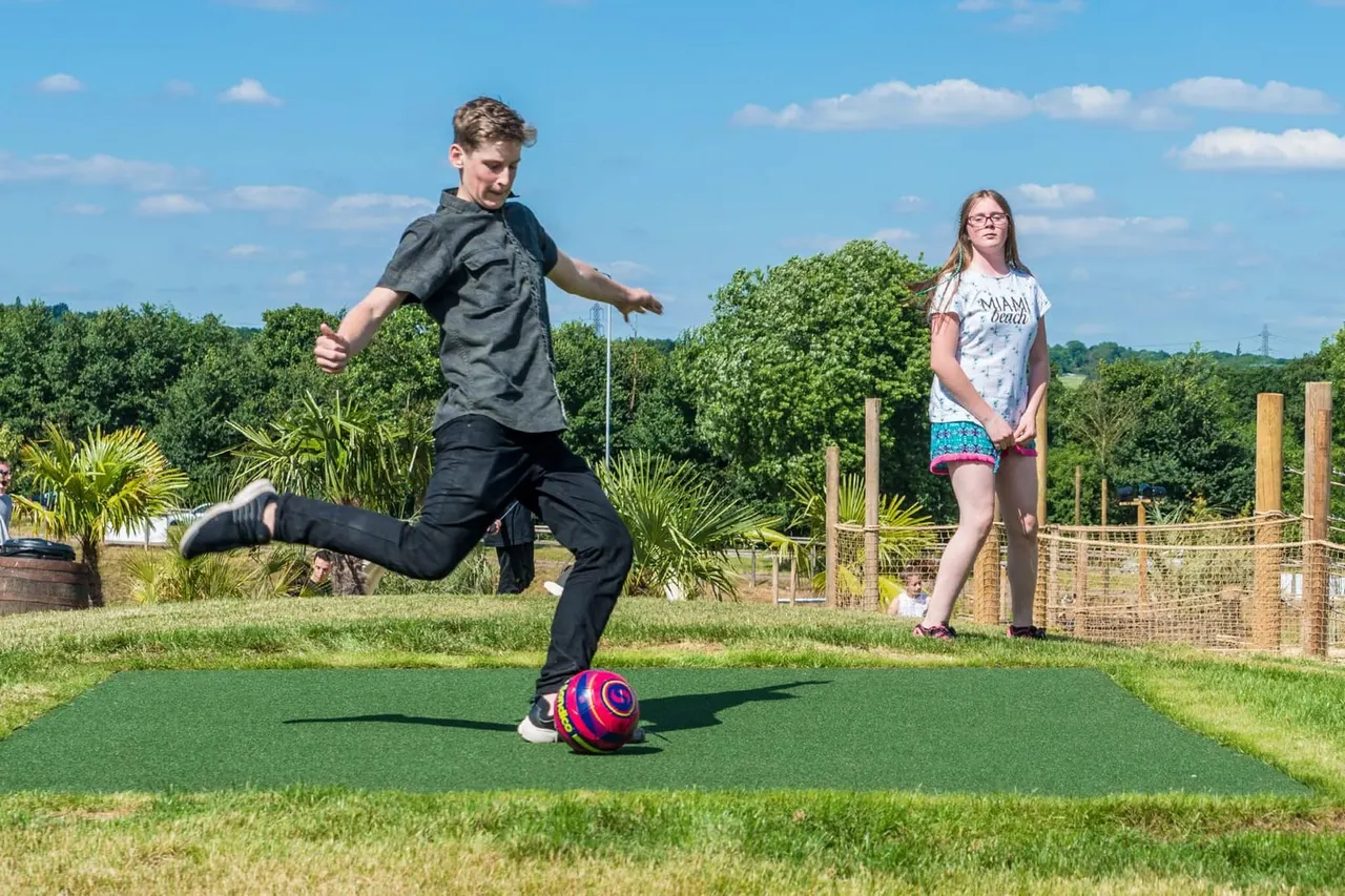 Boy kicking a colourful football on an adventure golf course whilst a girl watches, with tropical plants and wooden fencing behind.