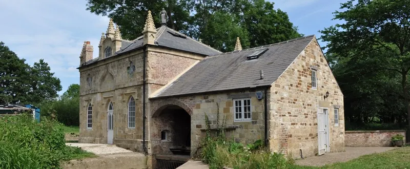 Historic stone mill building with arched entrance surrounded by mature trees and green grounds.