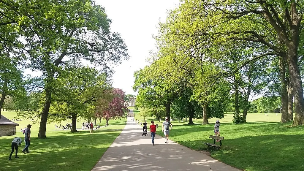 People jogging, walking and relaxing on a paved path through a large landscaped park with mature trees.