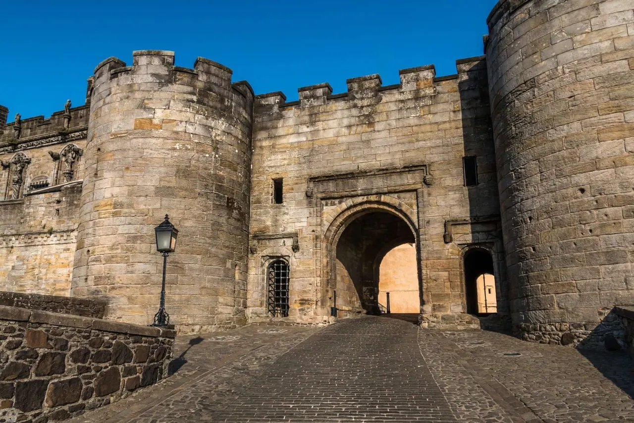 Stone castle with crenellated walls, towers and arched gateway under clear blue sky.