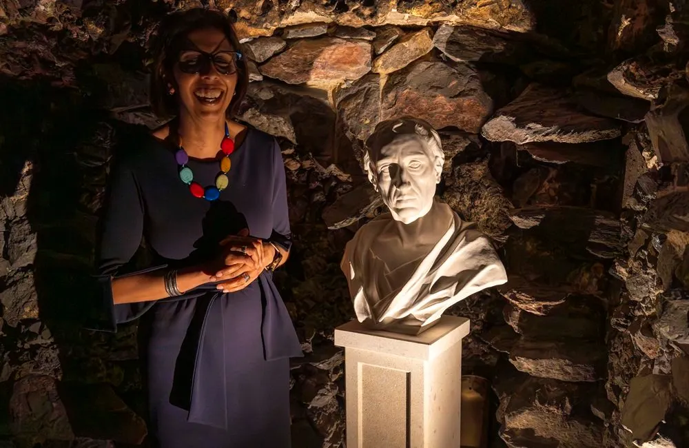 Woman in dark dress and colourful beaded necklace standing beside a stone bust displayed against a rustic stone wall.