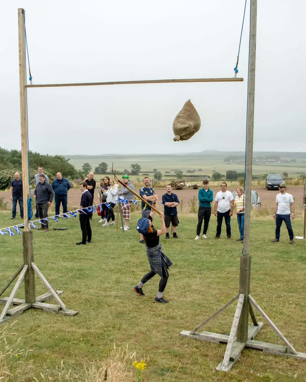 Participant in tartan competing in Highland games event, tossing large woven bag suspended from wooden frame, with spectators watching on grassy field.
