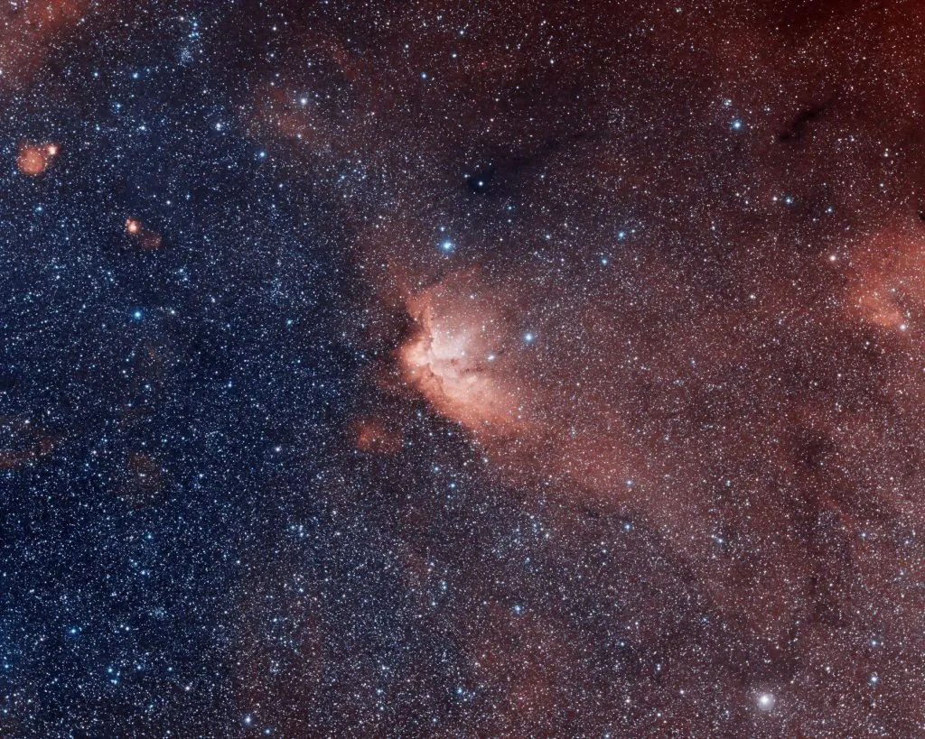 Deep space view showing countless stars across a dark sky with reddish nebula formations in the centre and blue stars on the left.