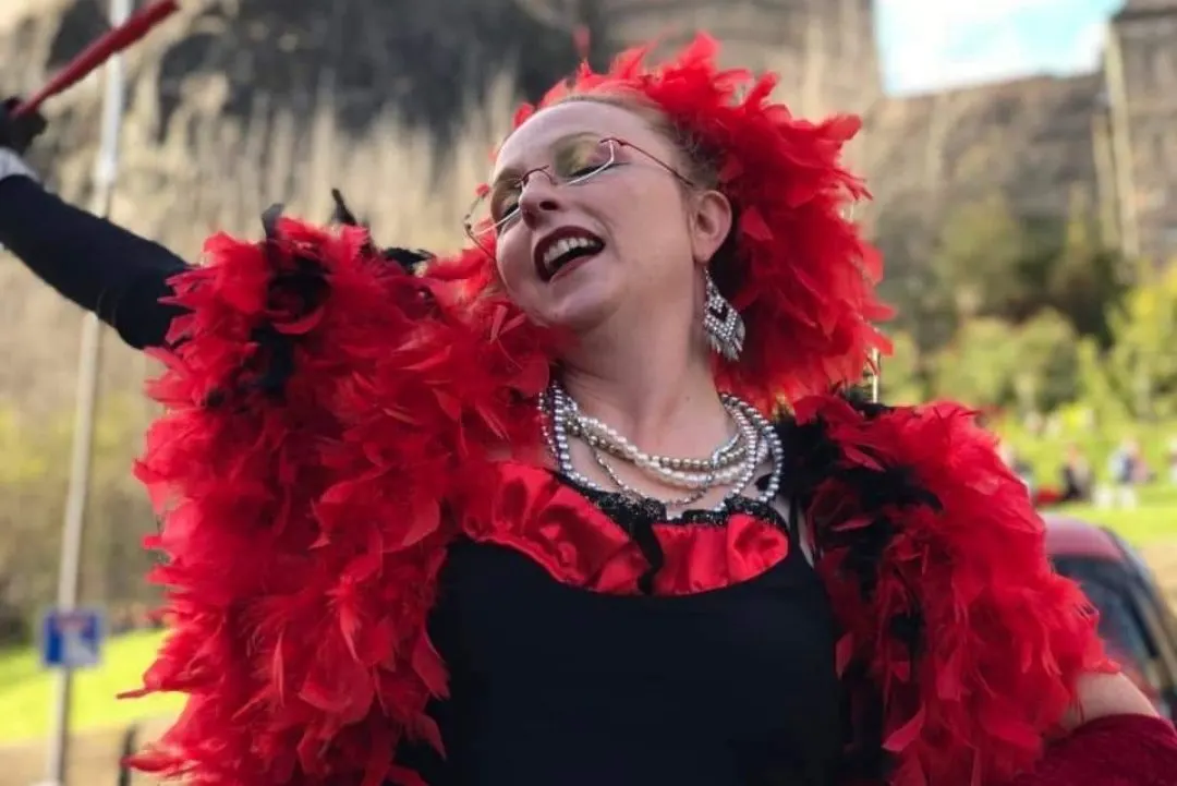 Performer in vibrant red feathered costume and red hair waves at audience in castle grounds during cabaret show.