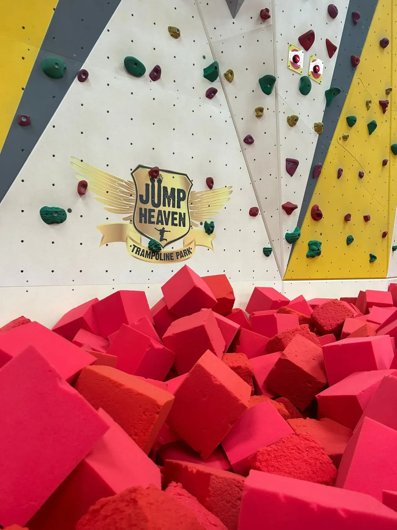 Indoor climbing walls with coloured holds above a bright pink foam pit at a trampoline park.