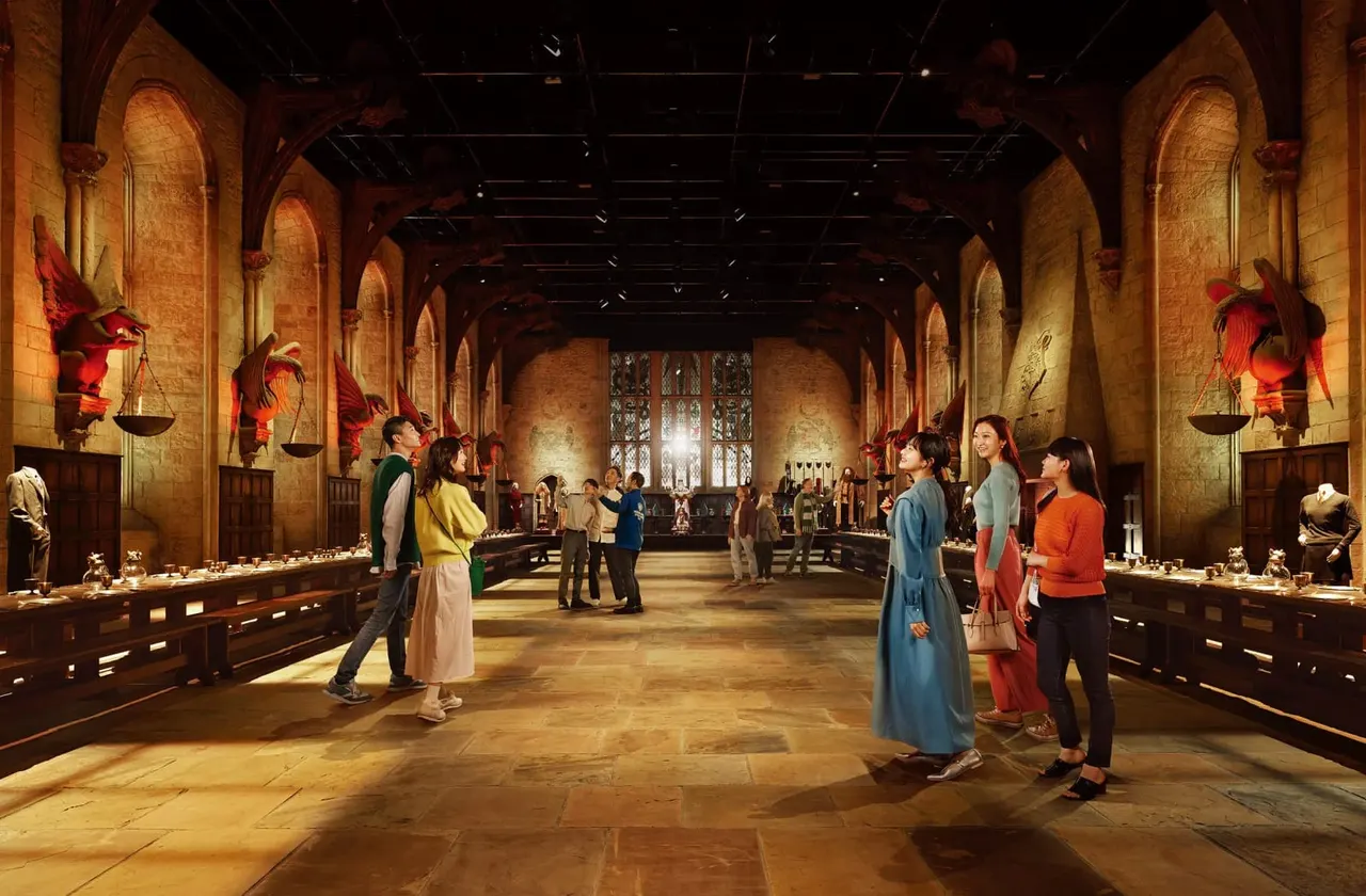 A themed interior set designed as a medieval Great Hall with stone arches, decorative red banners and large birds, wooden tables, and visitors exploring the space.