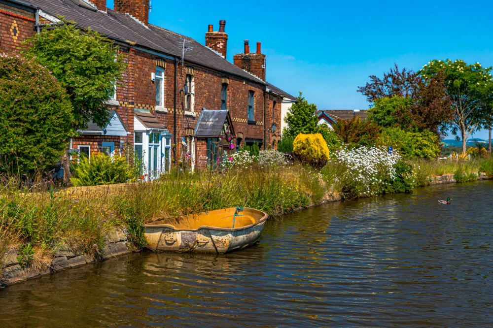 A yellow rowing boat moored beside brick cottages on a canal, with overgrown banks and gardens under clear blue sky.