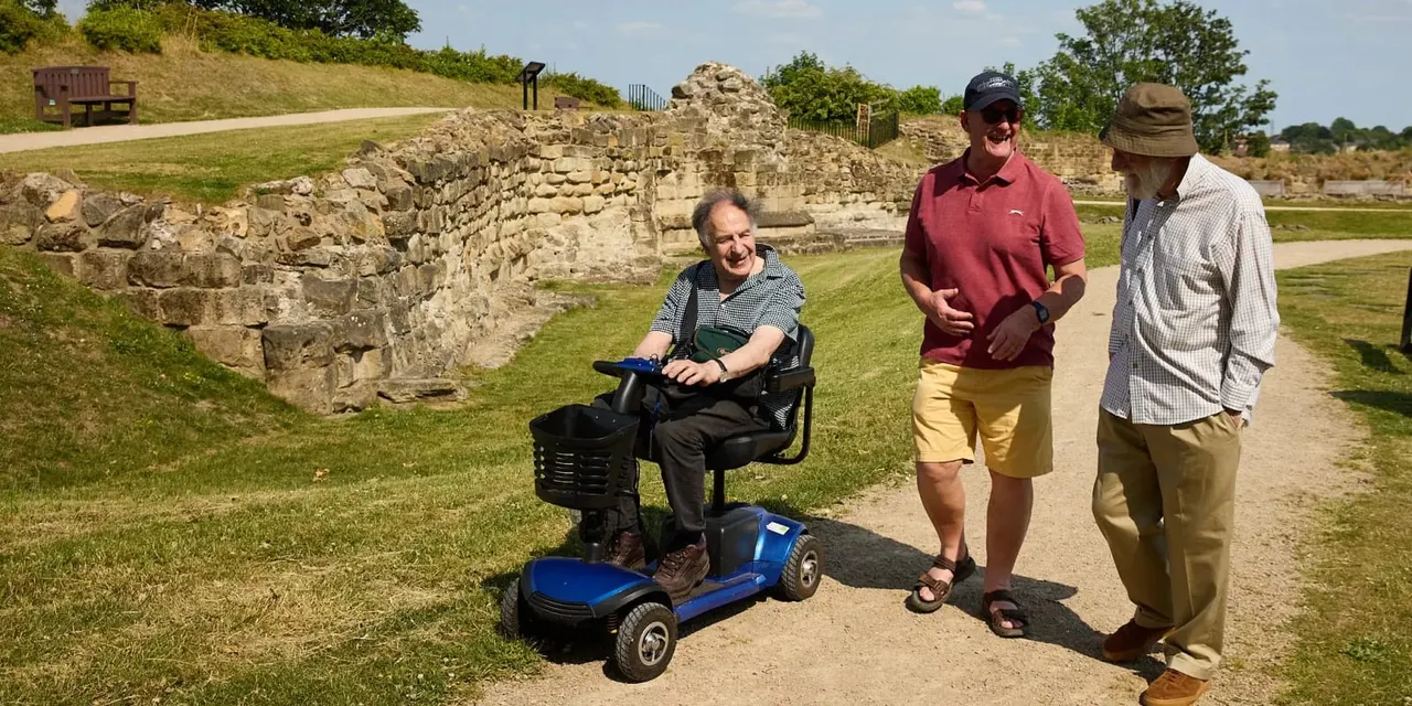 A man on a mobility scooter talks with two other visitors beside the ruins of a stone castle wall on a sunny day.