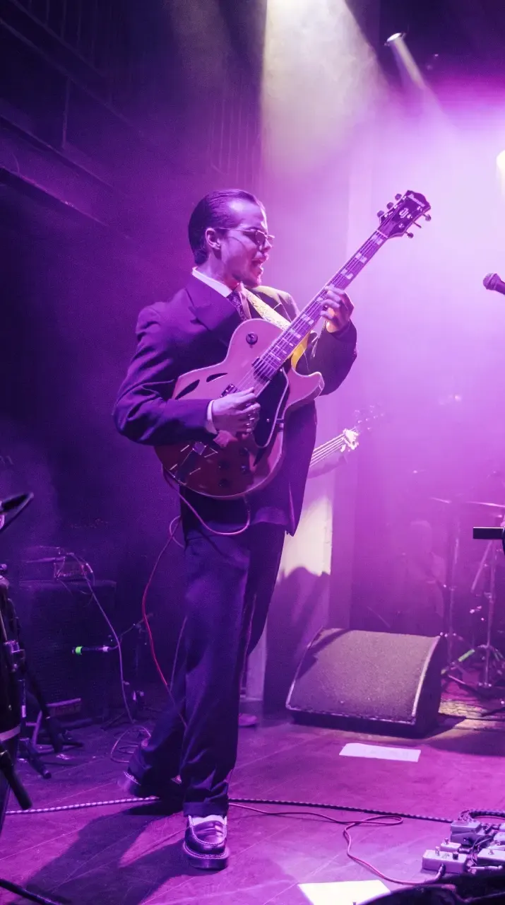 Musician in formal attire playing electric guitar on stage under purple stage lighting.