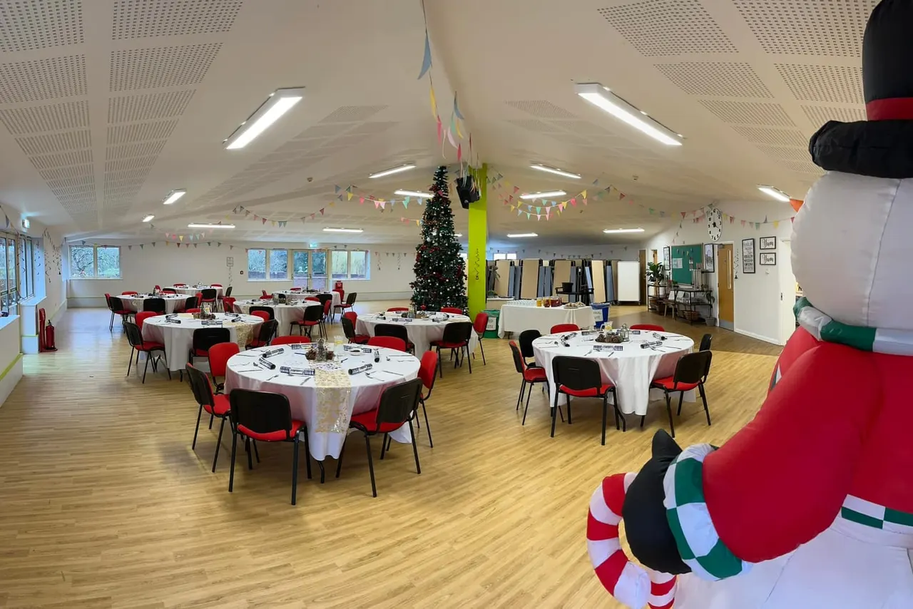 Spacious indoor hall decorated for Christmas with round tables set for dining, red chairs, festive bunting, and a decorated tree.