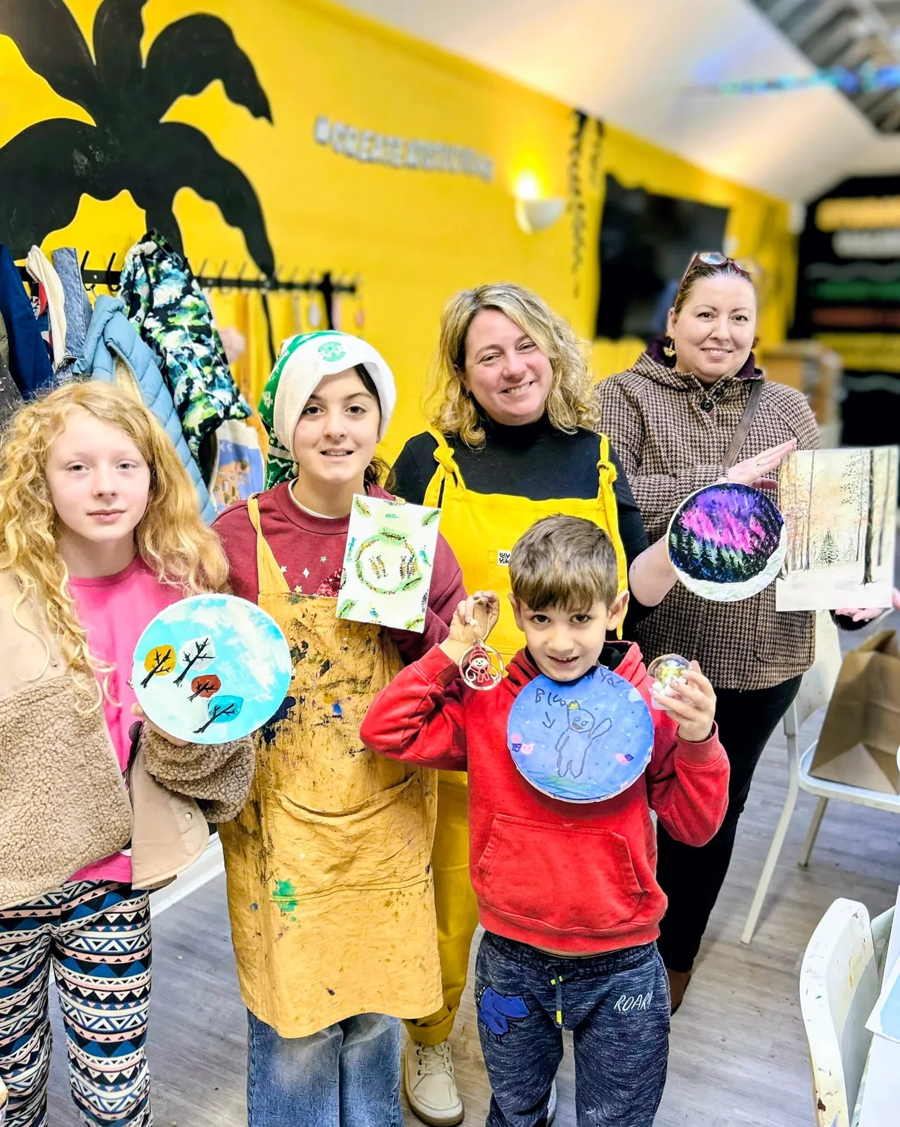 Children and adults holding hand-painted plates at an indoor creative workshop activity.