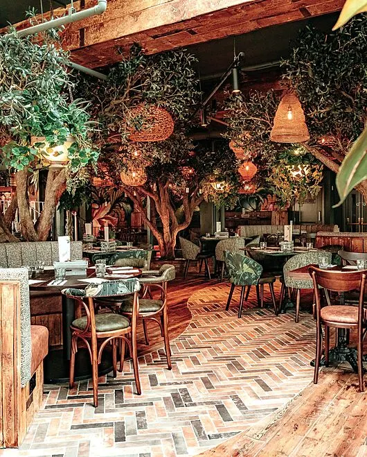 An indoor dining space with wooden furniture, hanging plants and pendant lights, herringbone flooring, and lush foliage throughout.