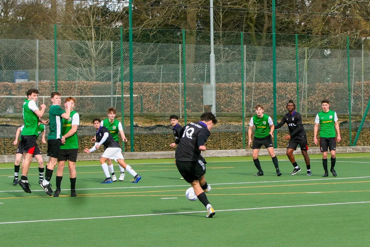 Young footballers in green and black kits playing on an artificial pitch enclosed by fencing.