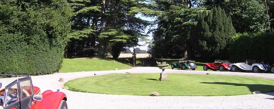Classic and vintage cars parked around a circular lawn at a country estate with mature trees.