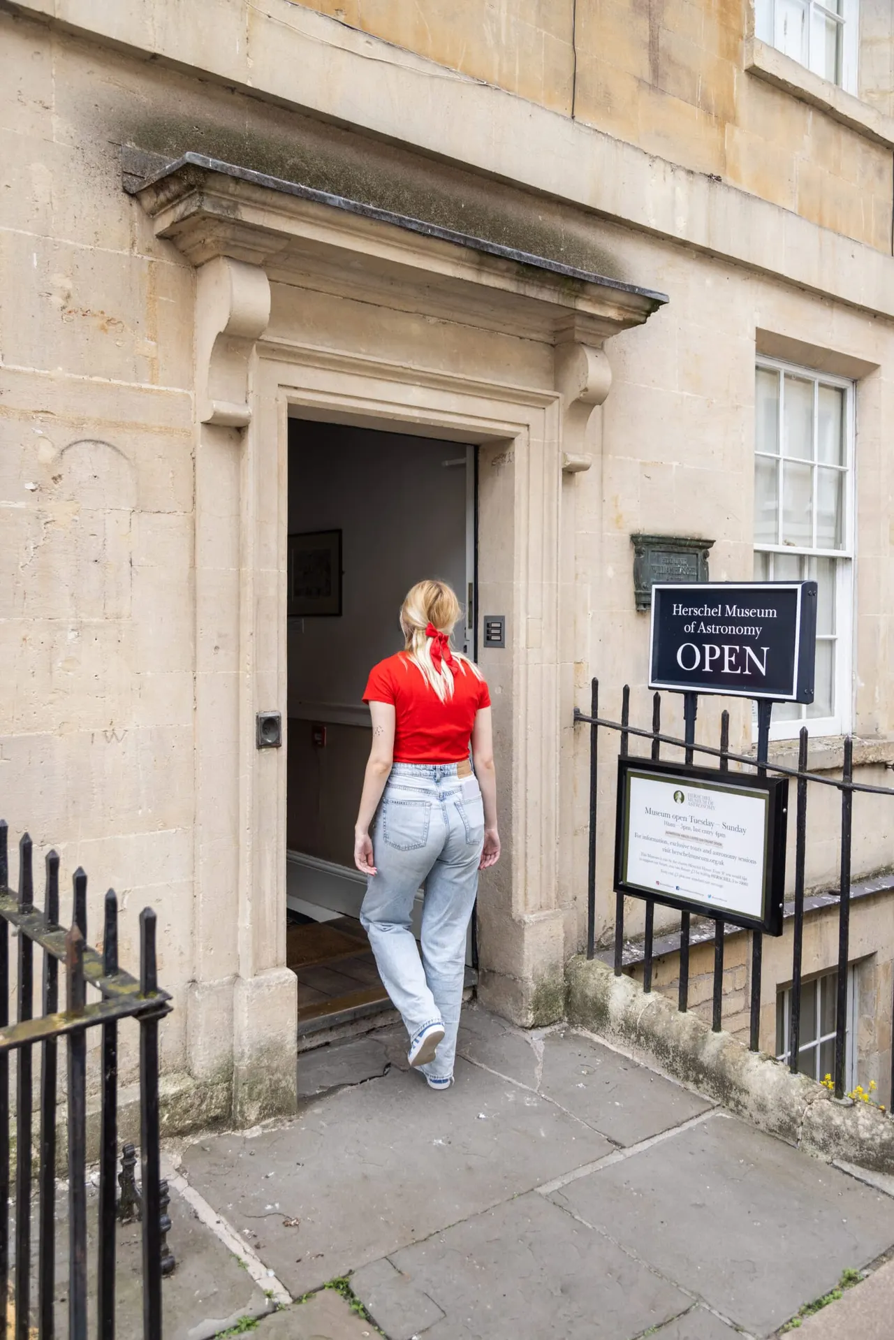 A woman in a red top and light jeans enters a stone Georgian building marked as the Herschel Museum of Astronomy, with an open sign visible.