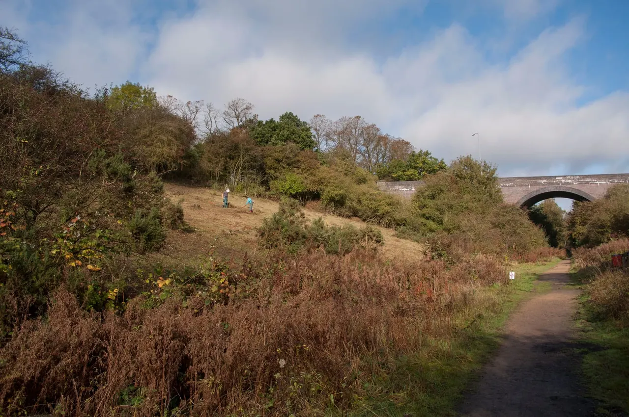 Two people walking on a hillside with autumn vegetation and a stone bridge visible in the distance.