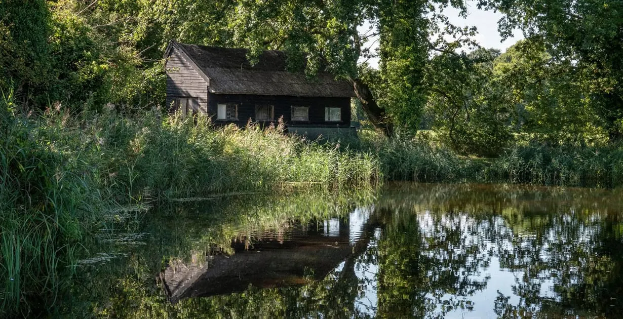 A historic thatched boathouse surrounded by dense reeds and trees beside a still waterway.
