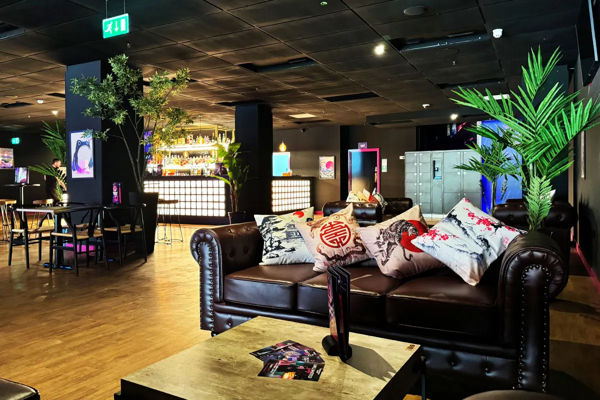 Modern indoor leisure space with black leather seating, colourful cushions, potted plants, bar area, and gaming screens.