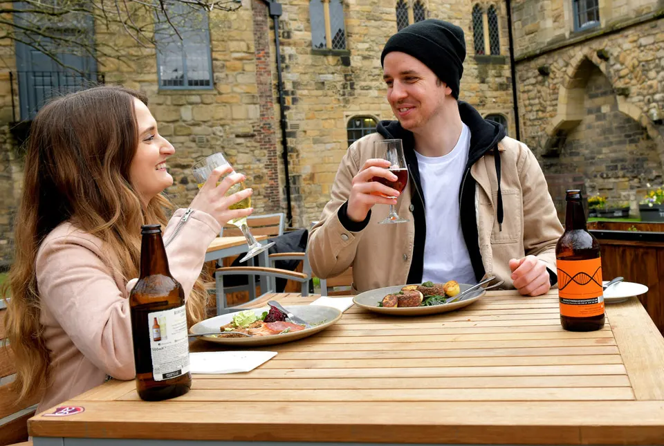 Two people toasting with drinks at an outdoor wooden table with food and beer bottles, stone building behind them.